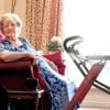Oudere vrouw zit in een stoel naast een rode binnenrollator met dienblad in een lichte woonkamer.