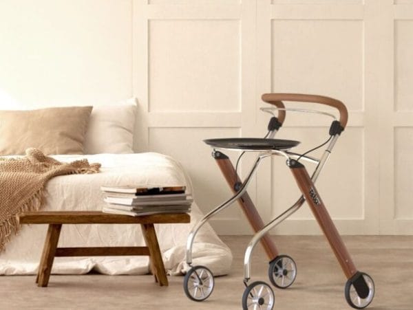 Binnenrollator Let's Go Indoor met dienblad in een lichte woonkamer naast een bed en houten bankje.