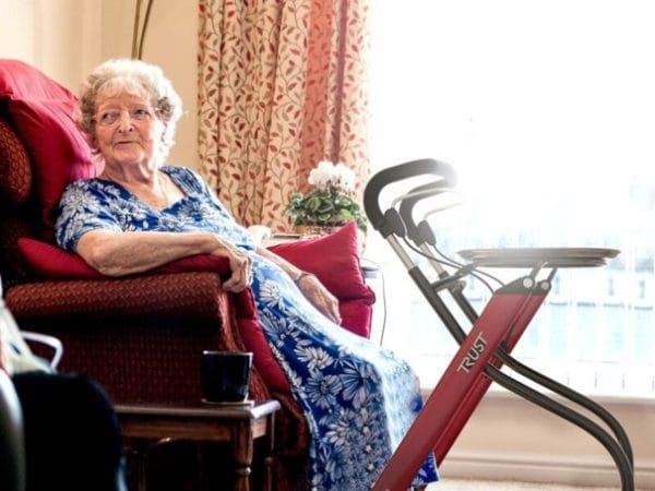 Oudere vrouw zit in een stoel naast een rode binnenrollator met dienblad in een lichte woonkamer.