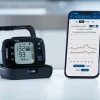 Omron RS7 Intelli IT pols bloeddrukmeter in houder naast smartphone met OMRON connect-app en bloeddrukgrafieken.