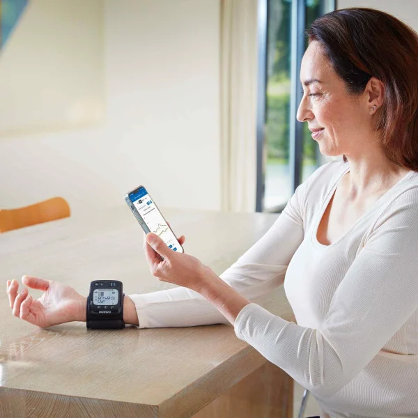 Vrouw meet polsbloeddruk met Omron RS7 Intelli IT en bekijkt de OMRON connect-app op haar smartphone aan de keukentafel.
