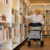 Rollator Rollz Air carbon rollator, zijaanzicht naast vrouw die in bibliotheek een boek pakt; met grijze zitting en tas.