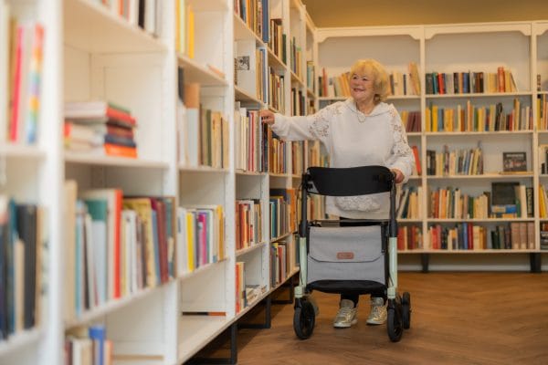 Rollator Rollz Air carbon rollator, zijaanzicht naast vrouw die in bibliotheek een boek pakt; met grijze zitting en tas.