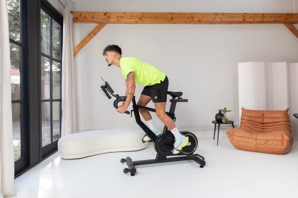 Man traint thuis op de VirtuFit ETAPPE 2.0i indoor cycle bike in een moderne lichte woonkamer met groot raam en designstoel.