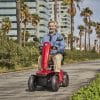 Man rijdt op een rode Vermeiren Sync scootmobiel over een pad met palmbomen en moderne gebouwen.