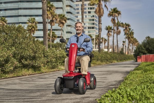 Man rijdt op een rode Vermeiren Sync scootmobiel over een pad met palmbomen en moderne gebouwen.