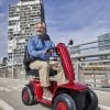 Man rijdt op een rode Vermeiren Sync scootmobiel over een stadsbrug met hoge gebouwen op de achtergrond.