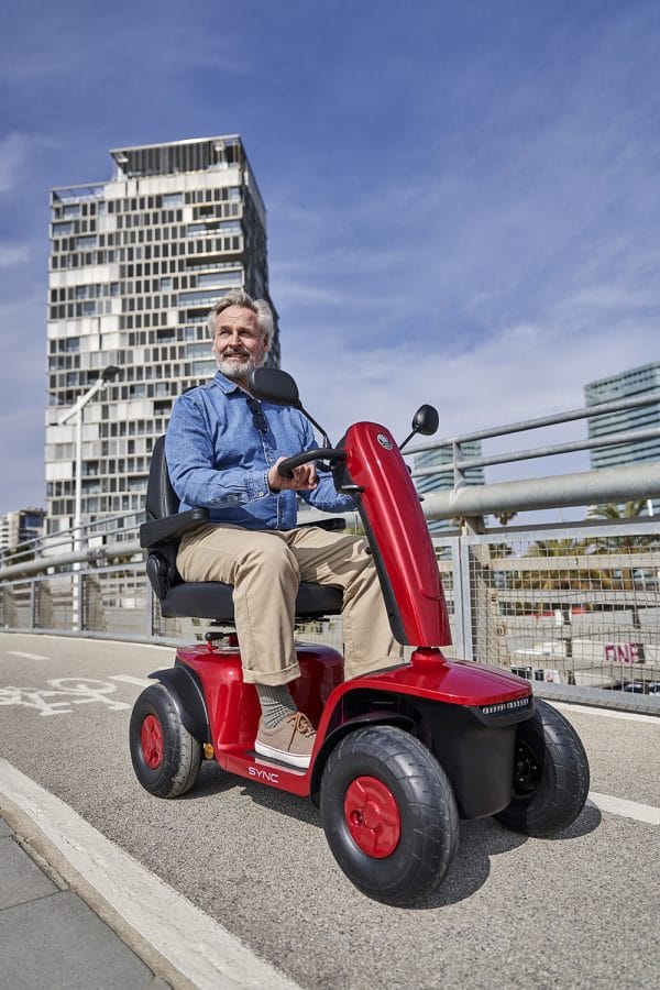Man rijdt op een rode Vermeiren Sync scootmobiel over een stadsbrug met hoge gebouwen op de achtergrond.