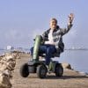 Man rijdt op de groene Vermeiren Sync scootmobiel over een pier langs rustig water en zwaait.