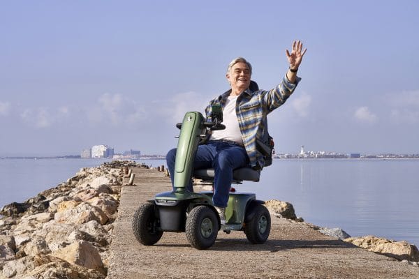 Man rijdt op de groene Vermeiren Sync scootmobiel over een pier langs rustig water en zwaait.