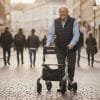 Oudere man loopt met de Rollator Rio met rugsteun en printaccenten over een geplaveide straat in de stad.