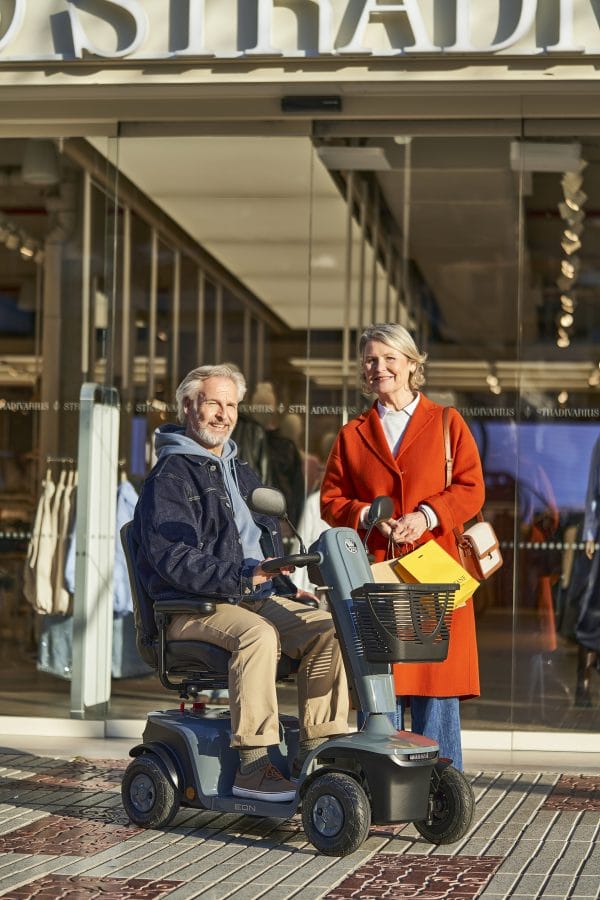 Man op de EON scootmobiel naast een vrouw met winkeltassen voor een winkelpand