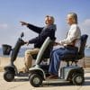 Twee volwassenen rijden op de Scootmobiel EON langs de kust, waarbij de bestuurder vooruit wijst.