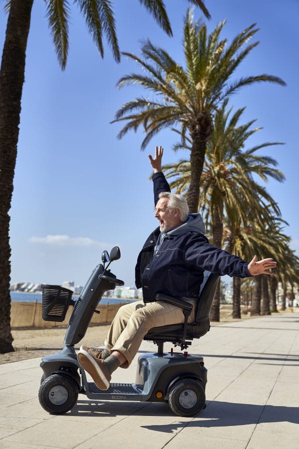 Man rijdt op de Scootmobiel EON over een boulevard met palmbomen en zee op de achtergrond