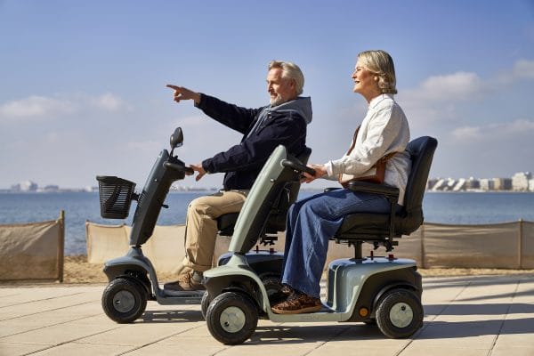 Twee volwassenen rijden op de Scootmobiel EON langs de kust, waarbij de bestuurder vooruit wijst.