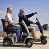 Twee volwassenen rijden op de Scootmobiel EON langs de kust, waarbij de bestuurder vooruit wijst.