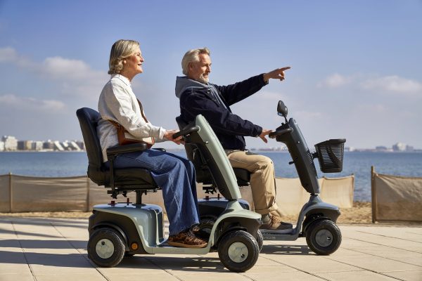 Twee volwassenen rijden op de Scootmobiel EON langs de kust, waarbij de bestuurder vooruit wijst.